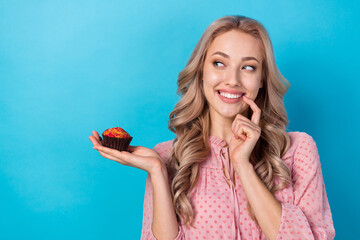 Poster - Photo of minded woman with wavy hairdo dressed pink clothes look at tasty muffin in arm finger teeth isolated on blue color background