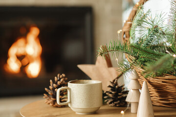 Wall Mural - Stylish cup of warm tea, wicker basket with fir branches, wooden trees and star, pine cones on table against burning fireplace. Modern christmas rustic eco friendly decor, winter hygge