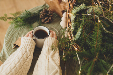 Wall Mural - Winter hygge. Hands in cozy sweater holding warm cup of warm tea on table with fir branches in rustic basket, wooden trees and star, pine cones and lights. Atmospheric cozy christmas time