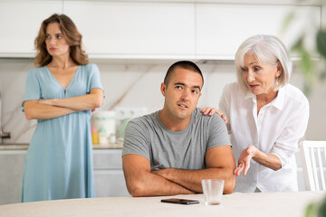 Wall Mural - Young wife and older woman quarrel son sitting at table