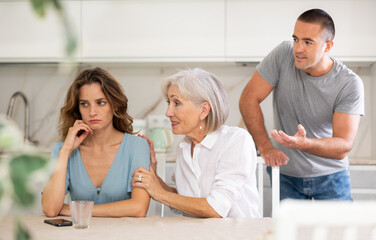 Wall Mural - Young girl is sitting at table in kitchen quarreling with her mother-in-law and husband