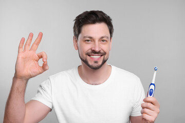 Wall Mural - Happy man holding electric toothbrush and showing ok gesture on light grey background