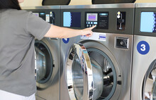 Young Woman Press The Button To Start The Washing Machine. Industrial Laundry Machines In Laundromat In A Public Laundromat. Self-service Laundry Facility With Many Automatic Washing Machines. Wash.
