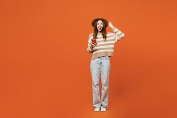 Wall Mural - Full body surprised excited young woman wear striped sweater hat casual clothes hold head use mobile cell phone isolated on plain orange red color wall background studio portrait. Lifestyle concept.