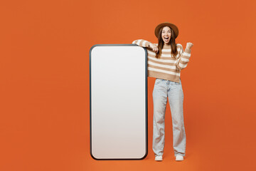 Sticker - Full body young woman she wearing striped sweater hat casual clothes big huge blank screen mobile cell phone smartphone with area do winner gesture isolated on plain orange red color wall background.