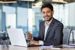 © Liubomir - Portrait of young hispanic businessman inside office at workplace, man holding bank credit card in hands, using laptop for online shopping and money transfer, employee smiling and looking at camera.