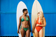 Portrait of a two female surfers standing in front of a blue background in swimwear with her surfboard by her side. Happy women doing beach activities during summer vacation.