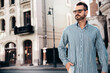 © halayalex - Portrait of handsome confident stylish hipster lambersexual model. Sexy modern man dressed in blue shirt and trousers. Fashion male posing on street background in Europe city at sunset. In sunglasses