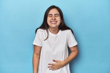 Young Caucasian woman on blue backdrop touches tummy, smiles gently, eating and satisfaction concept.