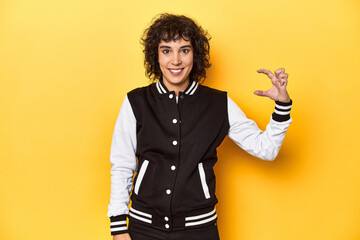 Curly-haired Caucasian woman in baseball jacket holding something little with forefingers, smiling and confident.