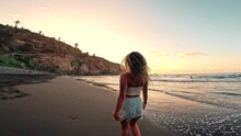 One Happy Young Girl Running And Walking Barefoot On The Sand Of The Beach With Sunset In The Background Enjoying And Having Fun At Summer. Blonde Woman Looking To The Sea Dancing And Jumping Lifestyl