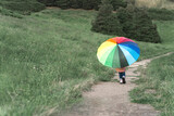 Fototapeta Tęcza - Child with a rainbow umbrella. Back view. A girl walks against a green background of nature. Childhood concept