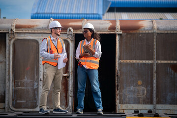 Engineers survey team wearing safety uniform and helmet under conversation document on hand and tablet inspect survey checking construction railway work station with oil refinery factory background.