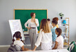 © Studio Romantic - Happy children at private school take turns answering tasks of young teacher near the blackboard. Beautiful young teacher attentively listens with interest to the answers of students in modern school.