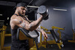 Bearded athlete in black vest and cap. He is lifting a dumbbell, training his biceps, sitting on preacher curl bench at dark gym. Close up