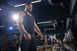 Tattooed, bearded athlete in black shorts, vest, cap. Going to do exercises with dumbbells, looking at set of black weights. Dark gym. Close up