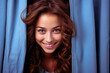 Charming young woman peeking timidly from behind a large blue curtain, embodying innocence and endearing vulnerability on a light blue background.