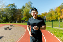 Portrait Of A Young Asian Sportsman Runner In Black Sportswear Running In The Stadium And Smiling At The Camera.