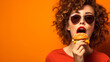 A cute girl appetizingly bites a burger, enjoying junk food, standing on an orange background.