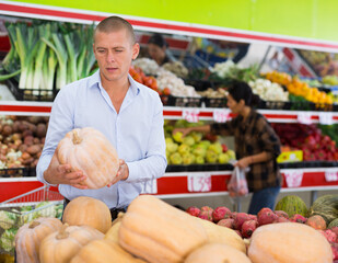 Sticker - European man standing in salesroom of greengrocer and choosing pumpkin. Woman selecting fruits in background.