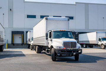 Maneuverable day cab middle duty rigs semi trucks with capacious box trailers standing on warehouse parking lot and in dock gates waiting for appropriate time for loading cargo for the next freight