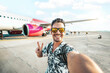 © Davide Angelini - Happy young man taking selfie picture with smart mobile phone boarding on air plane - Cheerful tourist standing at the airport - Travel life style concept with smiling guy going on summer vacation