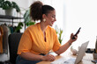 © Studio Marmellata - Portrait of content female fashion designer smiling and looking at mobile phone while working at table with laptop in atelier