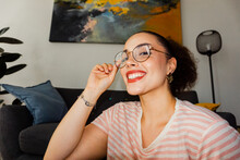 Positive young female in casual clothes and eyeglasses smiling with open mouth and looking at camera while sitting on rug floor beside sofa in living room