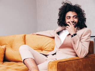 Handsome smiling hipster model. Unshaven Arabian man dressed in elegant beige suit . Fashion male with long curly hairstyle posing at home. sitting on yellow sofa