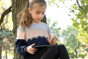 Wall Mural - Little girl uses a tablet while sitting in the park in the summer.