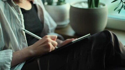 Female artist drawing on tablet. Creative woman holding notepad with digital pencil in hands and working remotely from home. Freelancer using computer to create art sitting near window