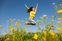 Happy And Beautiful Young Woman In A Bright Yellow Sweater And Blue Jeans  Jumping High In A Sunny Summer Field.