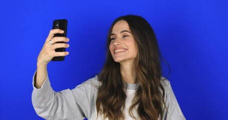 Wall Mural - Happy young woman blogger influencer holding modern smart phone wave hand hello. Smiling vlogger girl looking at mobile make video call, shooting vlog taking selfie on blue background, hi gesture.