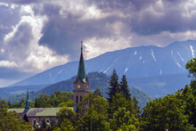 A Brick Church With A Green Spire And A Clock In The Foreground, Surrounded By Trees And Greenery, In Zakopane, Poland. Snowy Mountain Range And Cloudy Sky In The Background, Warm And Peaceful Mood