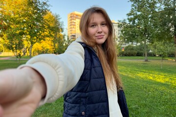 Portrait of happy cheerful young beautiful woman walking outdoors at summer or golden autumn park, smile and look at camera and give hand to her boyfriend