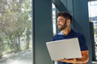 © Daria - Smiling male sales manager working on laptop standing in office during working day and looks away