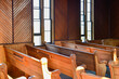 Interior of a vintage church with wooden pews or benches of red wood. The sun is shining in the building through tall clear glass windows. 