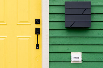 A vibrant yellow colored solid metal door with a black handle and deadbolt lock on a green wooden clapboard style house. There's a modern black square mailbox on the wall with an electrical box below.