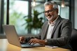 © Md Shahjahan - A smiling, busy mid-aged professional businessman works on his laptop at his office desk. An older, mature Indian businessman, a happy male entrepreneur worker, types on his computer