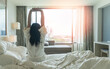 © Chinnapong - Hotel relaxation on lazy day with Asian woman waking up from good sleep on bed in weekend morning resting in comfort bedroom looking toward city view, having happy, work-life quality balance lifestyle