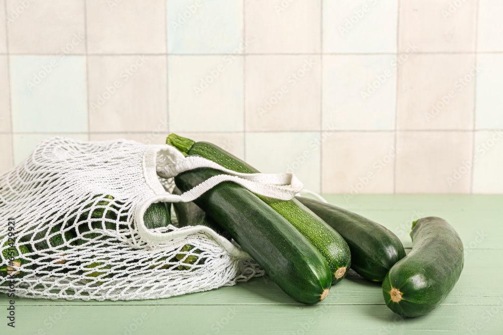 String bag with many fresh zucchini on green wooden table near white tile wall