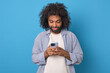 © antianti - Young positive curly Arabian man with phone in hands is typing SMS message or surfing internet and reading news in yellow press dressed in casual style stands posing on blue background.
