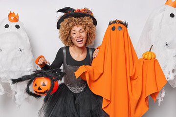 Wall Mural - Holiday celebration concept. Indoor photo of young cheerful pretty smiling African american witch holding pumpkin standing in centre surrounded by spooky orange and white ghosts at Halloween party