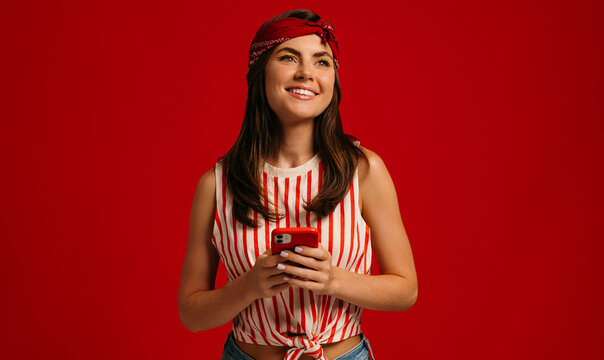 Stylish young hipster woman holding smart phone and smiling while standing on red background