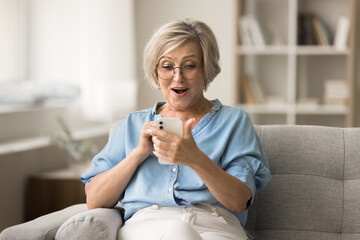 Surprised excited senior woman using mobile phone on home couch, staring at smartphone screen, reading message with good news, enjoying success, luck, fortune, shouting for joy