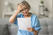 Stressed frustrated older woman getting bad news from paper letter, reading document at home, touching head in despair, panic attack, sitting on couch, receiving concerning medical result