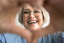 Cheerful pretty older mature woman in glasses looking at camera through finger heart shape, showing romantic symbol of love, kindness. Close up portrait of happy elder lady with perfect toothy smile