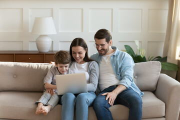 Wall Mural - Happy young family with cute little son using laptop at home together, smiling mother and father wearing glasses having fun with preschool child, looking at screen, watching cartoons, shopping online