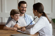 Female pediatrician wearing white coat checking smiling little patient heartbeat or lungs sounds, using stethoscope, cute preschool boy sitting on father laps at medical appointment, children checkup