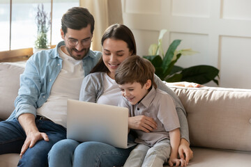 Wall Mural - Happy young parents with cute little son using laptop together, looking at screen, smiling mother and father wearing glasses with preschool child watching cartoons, shopping online, sitting on couch